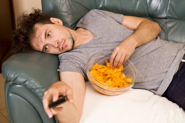 perezoso con el mando a distancia y patatas fritas en el sofá - pereza fotografías e imágenes de stock