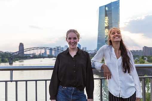 Two young women posing and smiling on city bridge over river with metropolis skyline in the background. Medium shot.