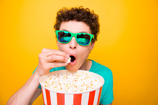Young attractive handsome funky guy wearing casual green polo t-shirt and 3d sun glasses, eating pop corn, watching cinema. Isolated over vivid shine bright yellow background