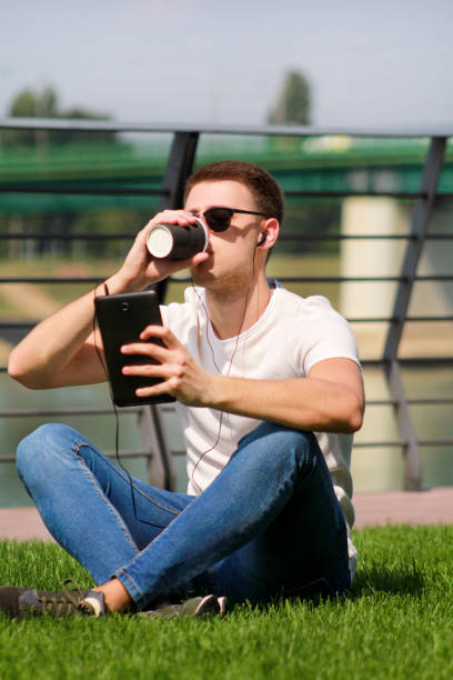 istock Young handsome guy with sunglasses using tablet and headphones, drinking coffee to go, man sitting on grass and enjoying nice sunny day by river side. Male model photo-shoot, urban fashion, lifestyle. 1038930422