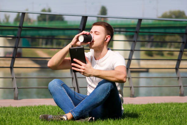 istock Handsome boy using tablet and headphones, drinking coffee to go, man sitting on grass and enjoying nice sunny day by river side. Model photo-shoot, urban fashion, lifestyle. White T-shirt and jeans. 1038930602
