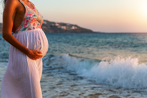 Pregnant woman in white summer dress stands on the beach and enjoys the sunset; closeup on belly