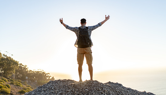 Rear view of young guy with backpack standing on the top of hill with arms spread open. Man enjoying the view from the top of the hill.