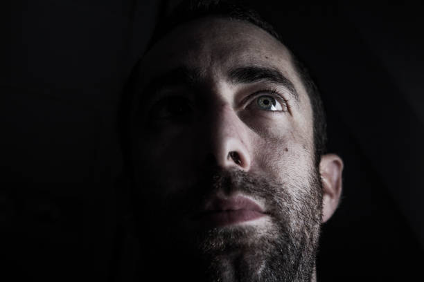 Dramatic portrait of a young man half expressionless face in deep shadow looking up isolated on black background Dramatic portrait of a young man half expressionless face in deep shadow looking up isolated on black background. attraction studio photography stock pictures, royalty-free photos & images