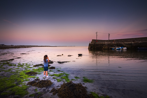 Barna is a coastal village in Connemara, west of Galway city in County Galway, Ireland. The little girl is 8 years old and is exploring the kelp filled beach  A dog is chasing fish in the shallow waters, with the beautiful sunset as a backdrop to the scene. She's calling her dog to return.