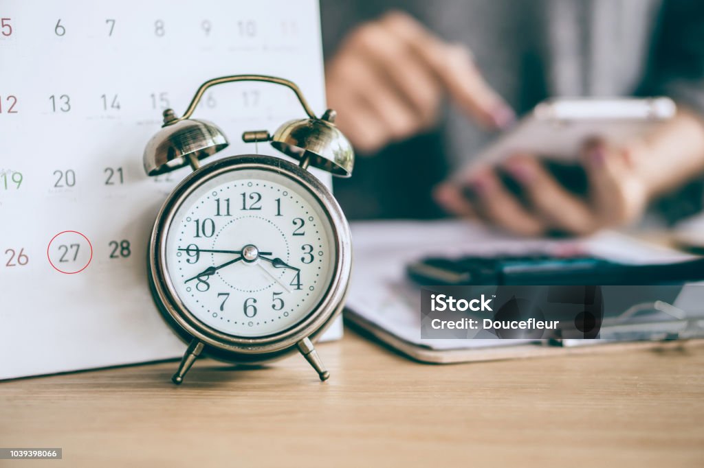 due date calendar and alarm clock with blur business woman hand calculating due date calendar and alarm clock with blur business woman hand calculating  monthly expenses during tax season. Time Stock Photo