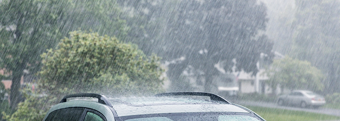 Drenched cars parked in residential home driveways during a torrential rain storm. It is raining cats and dogs, and the water is beating down and splashing so hard that it's difficult to see the neighbor's car and house just across the street through the low visibility fog. Suburban residential district near Rochester, New York, during an extreme weather mid-June summer downpour. Wide panoramic, cropped, 