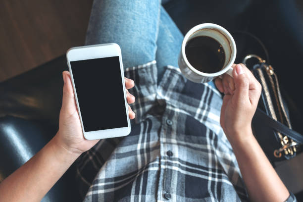 istock Top view mockup image of woman's hands holding white mobile phone with blank black desktop screen while drinking coffee in cafe 1039491268