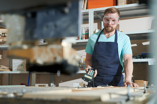 Portrait of young bearded carpenter standing with console and working on woodworking machine
