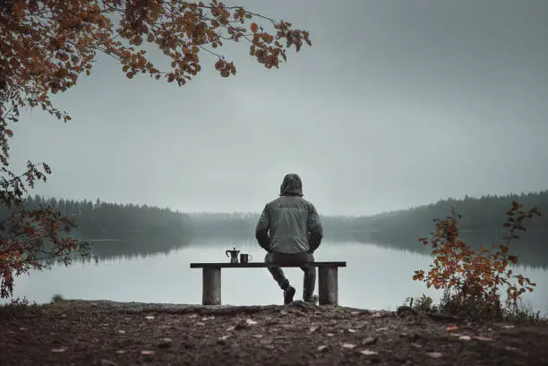 Photo of A man is sitting on a bench and looking at the lake. Back view. Also a coffee maker and a mug on the bench.