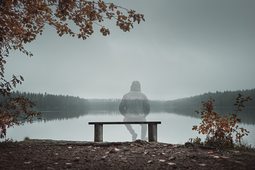A transparent man is sitting on a bench and looking at the lake. Back view. Autumn theme