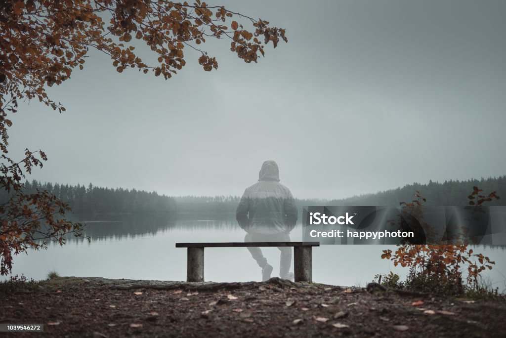 A transparent man is sitting on a bench and looking at the lake. Back view. Autumn theme A transparent man is sitting on a bench and looking at the lake. Back view. Foggy morning. Autumn theme Invisible Stock Photo