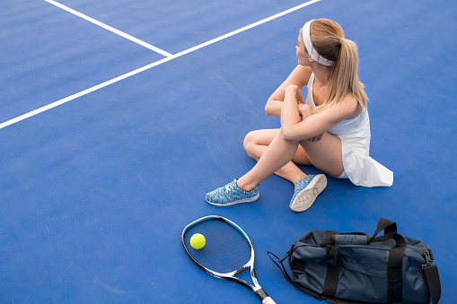 High angle  portrait of young blonde woman sitting on floor in indoor tennis court taking break from practice, copy space