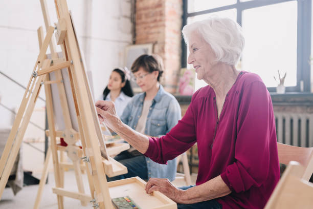 Senior Artist Painting in Art Studio Side view portrait of art students sitting in row and painting at easels in art studio, focus on smiling  senior woman enjoying work copy space hobbies stock pictures, royalty-free photos & images