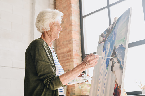 Side view portrait of white haired senior woman holding palette painting pictures at easel in  art studio standing against windows in sunlight, copy space
