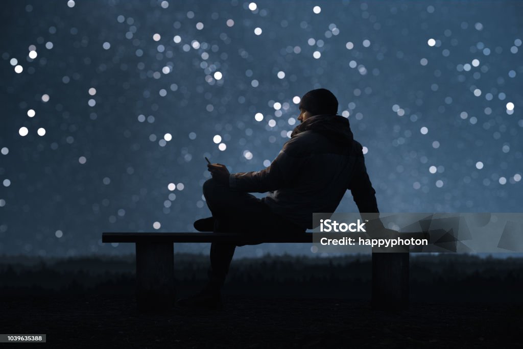 Ein Mann sitzt auf einer Bank und per Smartphone. Sternenhimmel Bokeh im Hintergrund - Lizenzfrei Himmel Stock-Foto