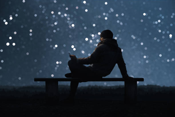 un hombre está sentado en un banco y con un smartphone. bokeh cielo estrellado en el fondo - ambiente atmosférico fotos fotografías e imágenes de stock