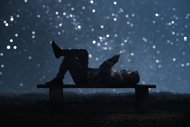 un hombre está tumbado en un banco y con un smartphone. bokeh cielo estrellado en el fondo - deshonestidad fotos fotografías e imágenes de stock