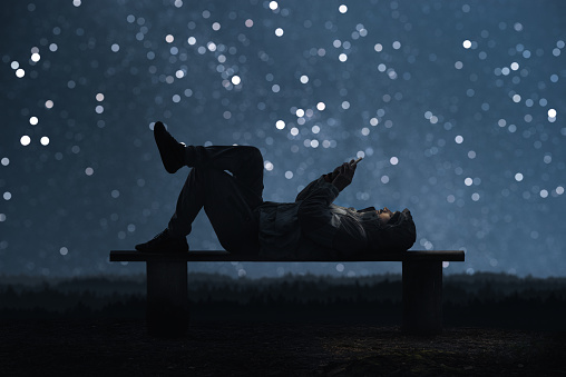 A man is lying on a bench and using a smartphone. Forest and starry sky bokeh In the background