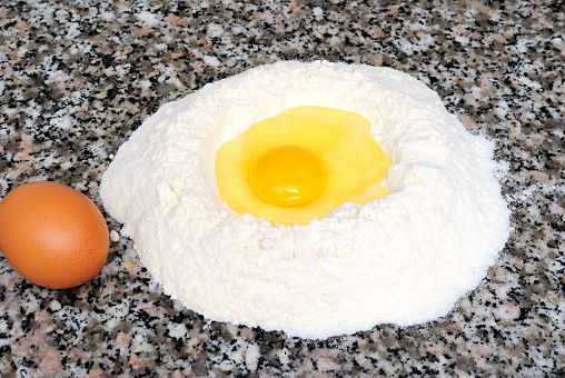 Flour fountain with an egg in the center, next to a whole egg, flat marble background, the ingredients to prepare desserts and home dough