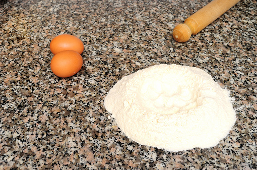 Flour fountain, two whole eggs and a rolling pin on the marble top, the ingredients to prepare desserts and dough at home
