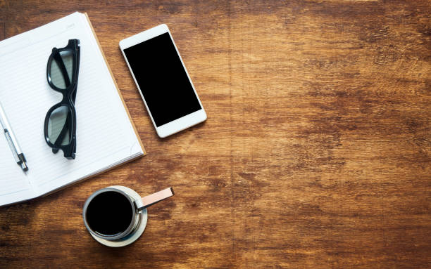 Wooden table with smartphone, eye glasses, open notebook, pen and coffee. Top view with copy space, flat lay. Studying concept