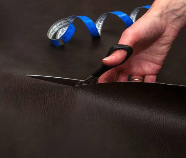 Woman hand cutting brown leather using scissors and blue twisted tape measure, close up. Sewing concept