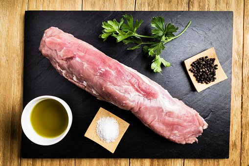 pork tenderloin next to salt, black pepper, a bowl of oil and parsley on a black slate plate on a wooden table