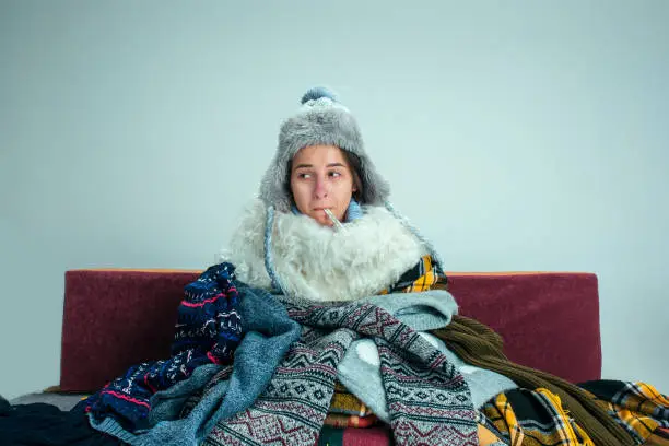 The young sick woman with flue sitting on sofa at home or studio covered with knitted warm clothes. Illness, influenza, pain concept. Relaxation at Home. Healthcare Concepts.