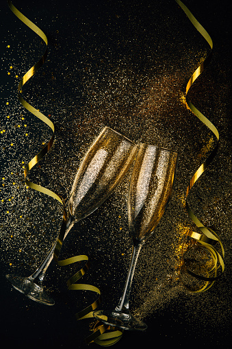 Two glasses of champagne toasting with golden confetti, glitter and serpentine on a dark background. Flat lay. Night of celebration concept