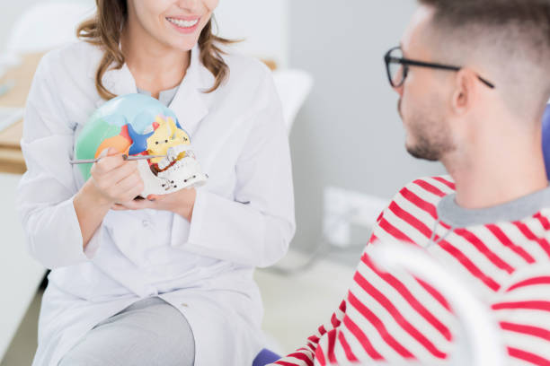 istock Close-up of smiling female dentist using dental mirror and model of skull while explaining tooth structure yo patient 1039922634