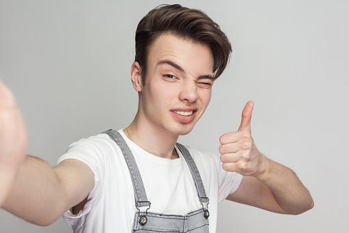 Selfie time! Portrait of happy handsome blogger teenager wearing in demin overalls standing, winking and thump up with toothy smile and making selfie. Indoor, isolated, studio shot, grey background