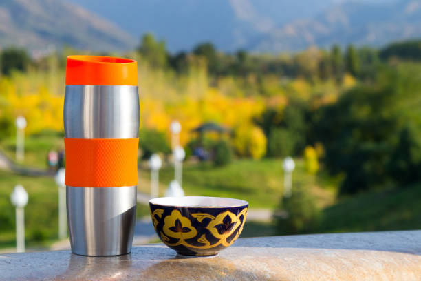 Closeup of a steel vacuum flask and a kazakh national cup. Blurred trees and mountains are in the background. View from a balcony, warm sunny september afternoon