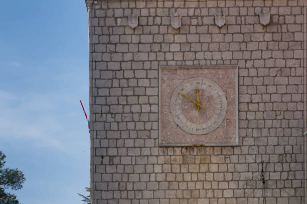 Tower made of stone tiles with clock. Clock are in a square, inner circle, roman numbers. Big square, historic part of town Krk, island Krk, Croatia.