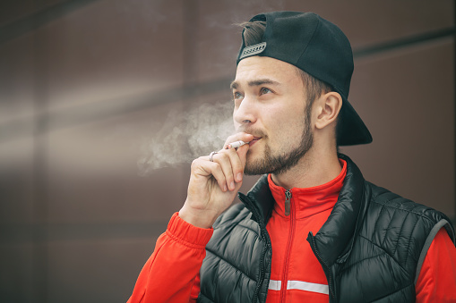 A young handsome white bearded man in black cap smokes a cigarette in the street in the autumn. Close up. Smoking activity.