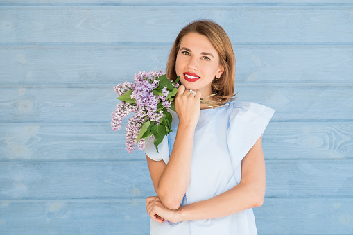 Beautiful portrait of young charming girl with bouquet of fresh lilac against blue wooden background