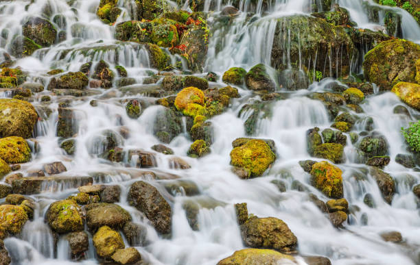 A close-up image showcasing a waterfall crashing down onto rocks and creating a frothy, turbulent pool below. The energy of the cascading water is evident, with droplets and foam dispersing dynamically upon impact. The contrast between the smooth water flow and the rugged rocks adds depth and texture to the composition.