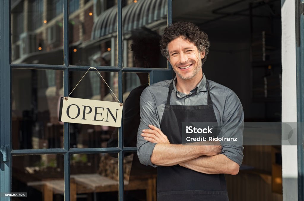 Owner standing outside restaurant Portrait of small business owner smiling and standing with crossed arms outside the cafe. Portrait of handsome smiling waiter standing in entrance of coffee shop. Successful mature man with arms crossed smiling at camera. Owner Stock Photo
