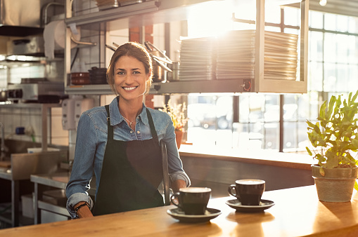 Smiling waitress wearing black apron standing behind counter in cafeteria and looking at camera. Mature woman serving two cups of hot coffee to customer in cafeteria. Small business and entrepreneur concept.