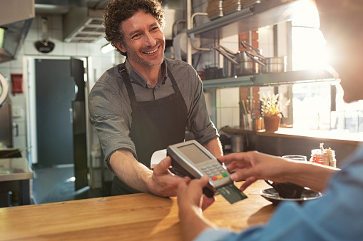 Woman paying by credit card and entering pin code on reader holded by smiling barista in cafeteria. Customer using credit card for payment. Mature cashier wearing apron accepting payment over nfc technology.