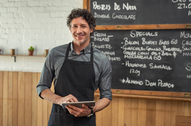 Waiter holding digital tablet at cafeteria Mature waiter wearing black apron and standing in front of the blackboard with the menu of the day. Portrait of smiling man holding digital tablet and looking at camera. Happy small business owner working in cafeteria with digital tablet. cafe tablet stock pictures, royalty-free photos & images