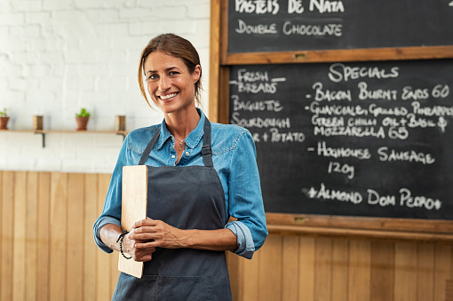 Portrait of smiling mid owner holding clipboard in hand while looking at camera. Beautiful waitress at cafeteria wearing black apron welcoming customers. Mature woman working at small business shop.