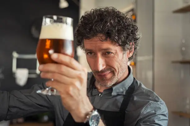 Mature man examining the quality of craft beer at brewery. Inspector working at alcohol manufacturing factory checking beer. Man in distillery checking quality control of draught beer.