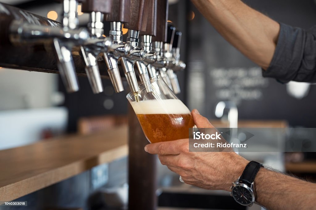 Pouring beer in glass Hand of bartender pouring a large lager beer in tap. Closeup of hand serving beer in glass using tap. Close up of barman hand at beer tap pouring an amber draught beer at pub. Beer - Alcohol Stock Photo
