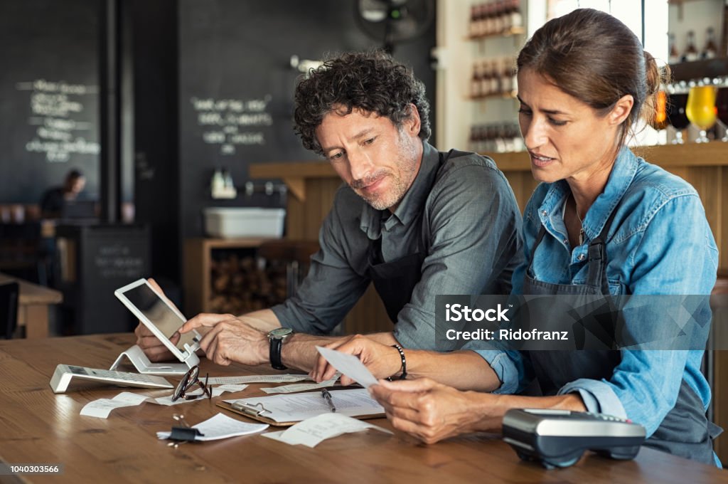 Staff calculating restaurant bill Man and woman sitting in cafeteria discussing finance for the month. Stressed couple looking at bills sitting in restaurant wearing uniform apron. Café staff sitting together looking at expenses and bills. Small Business Stock Photo