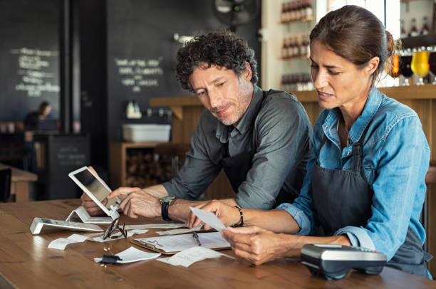 Staff calculating restaurant bill Man and woman sitting in cafeteria discussing finance for the month. Stressed couple looking at bills sitting in restaurant wearing uniform apron. Café staff sitting together looking at expenses and bills. ipad calculator stock pictures, royalty-free photos & images