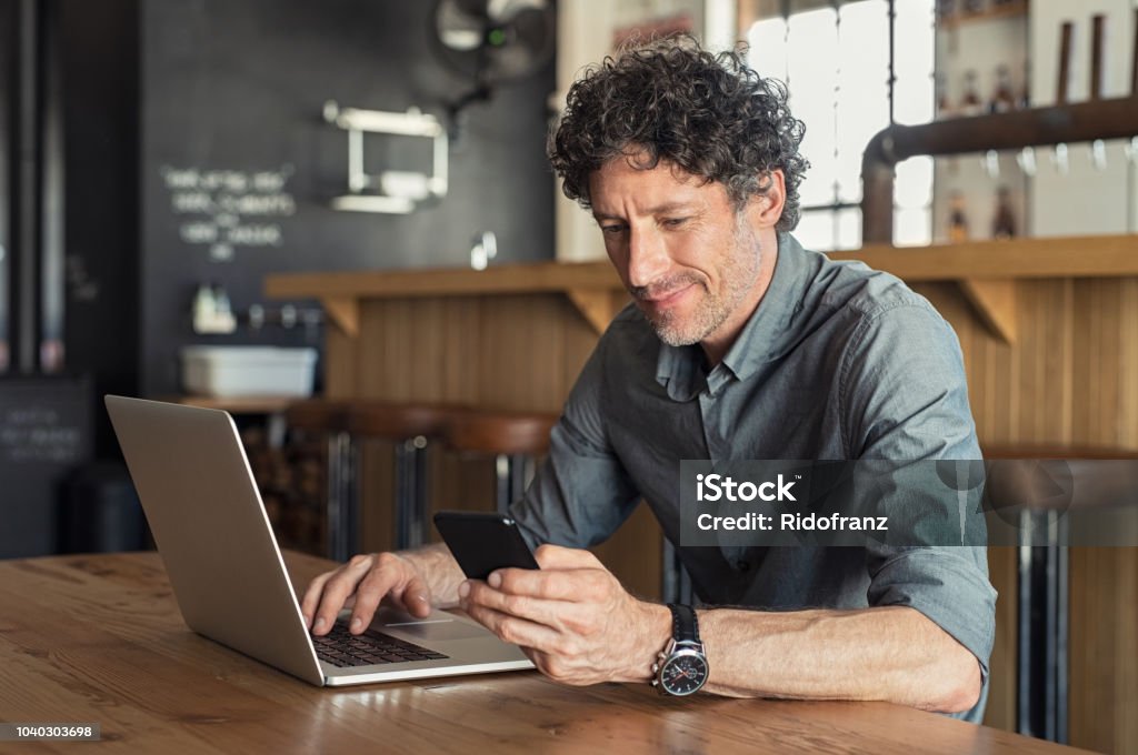 Mature businessman working at cafe Happy mature business man sitting at cafeteria with laptop and smartphone. Businessman texting on smart phone while sitting in a pub restaurant. Portrait of senior formal man working and checking email on computer. Men Stock Photo