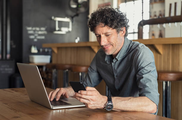 Mature businessman working at cafe Happy mature business man sitting at cafeteria with laptop and smartphone. Businessman texting on smart phone while sitting in a pub restaurant. Portrait of senior formal man working and checking email on computer. busy restaurant tables stock pictures, royalty-free photos & images