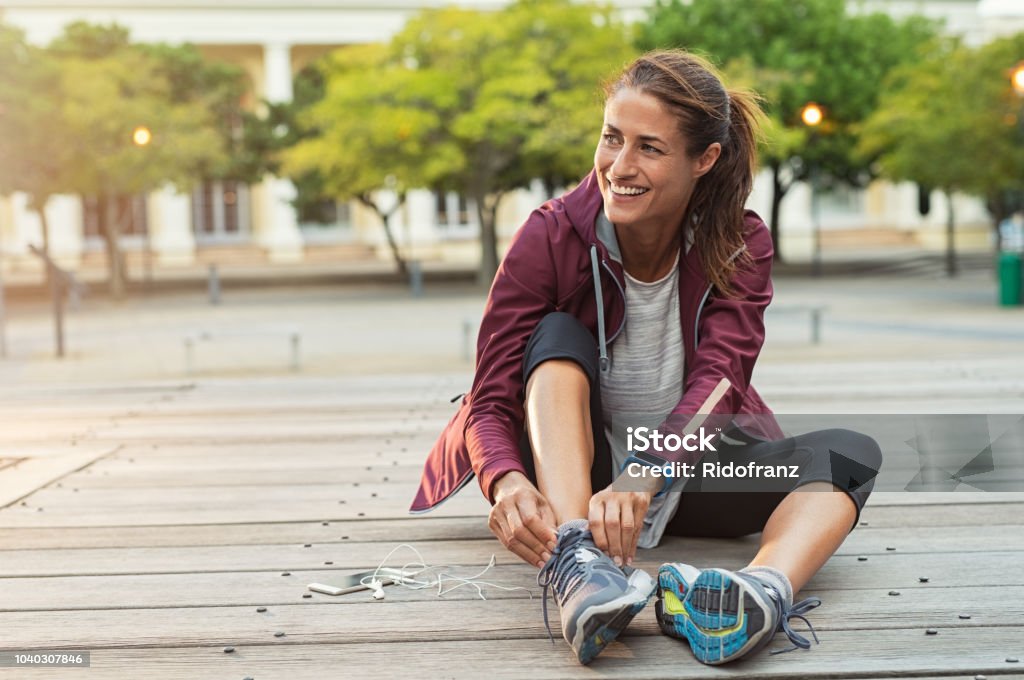 Woman wearing sport shoes Mature fitness woman tie shoelaces on road. Cheerful runner sitting on floor on city streets with mobile and earphones wearing sport shoes. Active latin woman tying shoe lace before running. Women Stock Photo