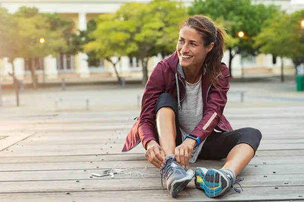 Mature fitness woman tie shoelaces on road. Cheerful runner sitting on floor on city streets with mobile and earphones wearing sport shoes. Active latin woman tying shoe lace before running.